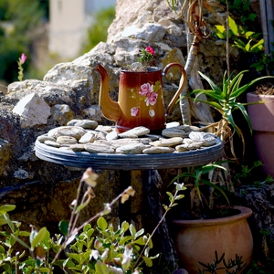 Table ronde surmontée de gallets et d'une cruche pot de fleur devant un mur de pierres et entourée de pots de plantes - France  - collection de photos clin d'oeil, catégorie clindoeil